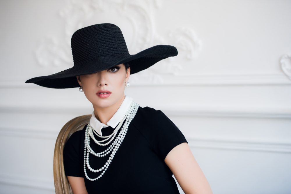 Attractive woman wearing black dress, hat and pearls, sitting on chair