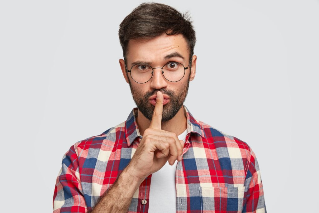 confident young bearded male makes shh gesture, keeps index finger over mouth and lifts eyebrow, asks for silence, cannot concentrate, isolated over white background. people and hush concept