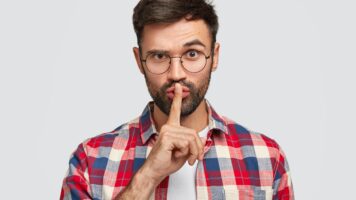 confident young bearded male makes shh gesture, keeps index finger over mouth and lifts eyebrow, asks for silence, cannot concentrate, isolated over white background. people and hush concept