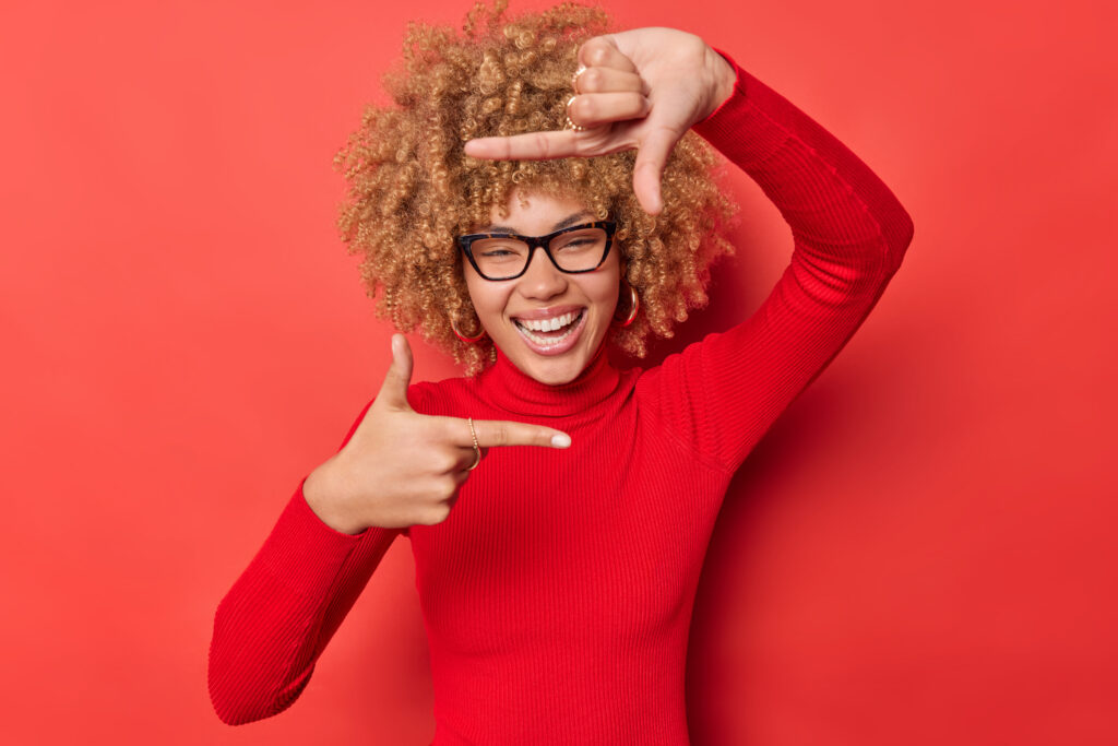 positive curly haired woman searches perfect angle takes photo makes frame gesture as if photographing with camera smiles positively wears spectacles and turtleneck isolated on red studio wall