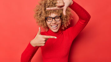 positive curly haired woman searches perfect angle takes photo makes frame gesture as if photographing with camera smiles positively wears spectacles and turtleneck isolated on red studio wall